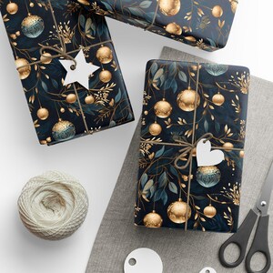 May include: Three wrapped gifts with a dark blue background featuring gold ornaments and green leaves. Each gift is tied with twine and a white tag. A ball of twine, scissors, and a tag are also visible.