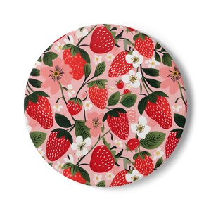 May include: Round decorative item with a pink background featuring a repeating pattern of red strawberries, green leaves, white flowers, and pink flowers. The strawberries have white seeds and green stems. The overall design is a floral and fruit pattern.