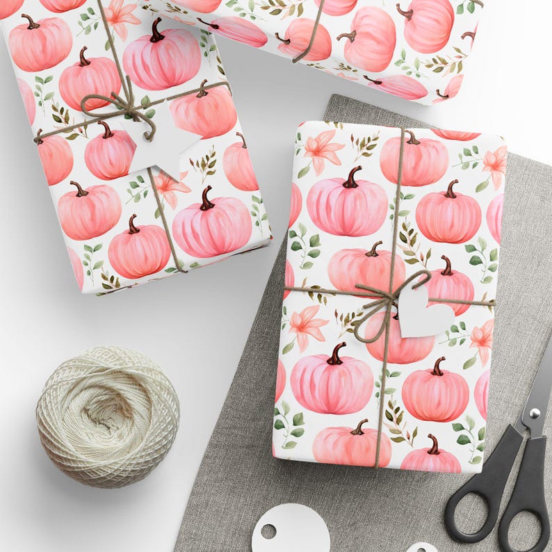 May include: Three gift boxes wrapped in white paper with a pink pumpkin pattern. The boxes are tied with twine and have white gift tags. The boxes are on a white surface with a spool of twine and a pair of scissors.