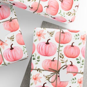 May include: Pink and white wrapping paper with a pattern of pink pumpkins and green leaves. The paper is wrapped around a gift box with a brown twine bow and a white heart-shaped tag.