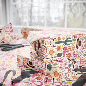 May include: Two gift boxes wrapped in colourful patterned paper with a Western theme. The paper features cacti, horseshoes, stars, and other Western motifs. The boxes are stacked on top of each other.