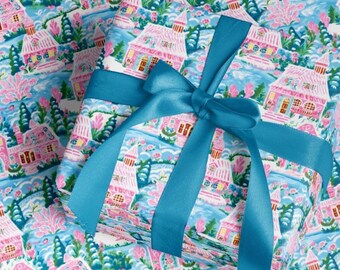 Könnte beinhalten: Geschenkpapier mit einer skurrilen Dorflandschaft in Rosa, Blau und Grün, gebunden mit einem türkisfarbenen Satinband. Das Geschenkpapier hat ein sich wiederholendes Muster aus Häusern, Bäumen und Wasser.