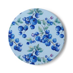 May include: A round, light blue coaster featuring a watercolor design of blueberries and green leaves on branches. The blueberries are various shades of blue and purple. The coaster has a white border and is set against a white background.