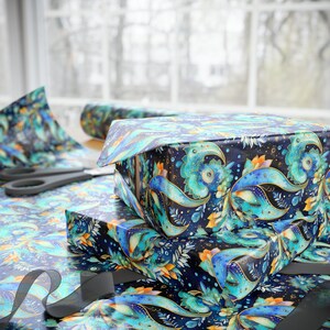 May include: Two gift boxes wrapped in patterned paper with teal, blue, and gold floral designs. The boxes are stacked on a table with wrapping paper, scissors, and a black ribbon. The background includes a window with a blurred view.