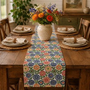 May include: A long rectangular table runner featuring a repeating pattern of stylized circular flowers in shades of red, orange, yellow, blue, and green on a white background, centered on a wooden dining table with place settings and a flower vase.