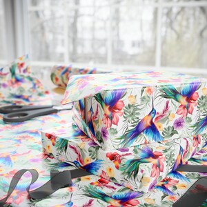 May include: A stack of gift boxes wrapped in white paper with a colorful hummingbird and floral pattern. The boxes are tied with a black ribbon.