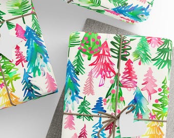 May include: Gift-wrapped presents with colorful watercolor Christmas tree patterns. The gifts are wrapped in white paper with a repeating design of green, blue, pink, and yellow trees. Each package is tied with twine and a white heart-shaped tag.