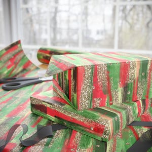Puede incluir: Cajas de regalo envueltas con papel de regalo con estampado de pinceladas rojas, verdes y doradas con purpurina. Unas tijeras negras y un rollo de papel de regalo est&aacute;n sobre la mesa. Una cinta negra est&aacute; en primer plano.