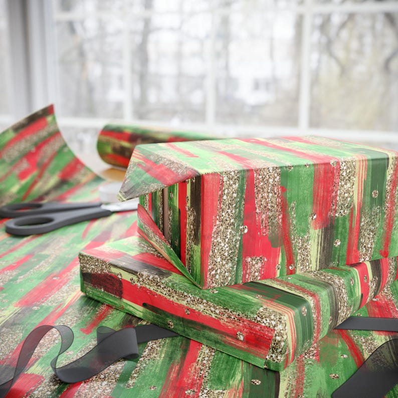 Puede incluir: Cajas de regalo envueltas con papel de regalo con estampado de pinceladas rojas, verdes y doradas con purpurina. Unas tijeras negras y un rollo de papel de regalo est&aacute;n sobre la mesa. Una cinta negra est&aacute; en primer plano.