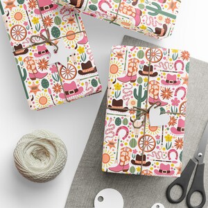 May include: Three gift boxes wrapped in white paper with a pink and yellow cowboy themed pattern. The pattern includes cowboy boots, hats, cacti, horseshoes, and suns. The boxes are tied with twine and have white gift tags attached.