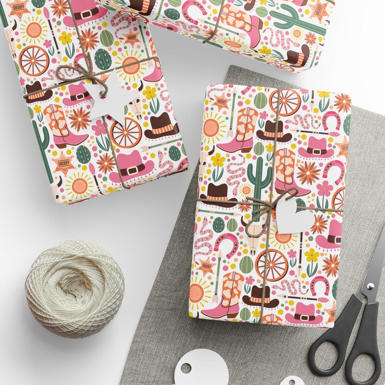 May include: Three gift boxes wrapped in white paper with a pink and yellow cowboy themed pattern. The pattern includes cowboy boots, hats, cacti, horseshoes, and suns. The boxes are tied with twine and have white gift tags attached.