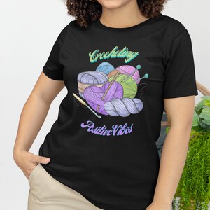 May include: A black t-shirt featuring a graphic of balls of yarn and crochet hooks. The text "Crocheting Positive Vibes" is printed on the shirt.