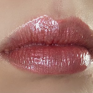 May include: Close-up of a person's lips wearing a glossy, shimmery, rose-gold colored lipstick.
