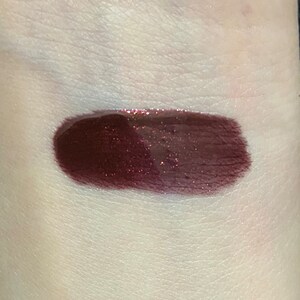 May include: A swatch of dark burgundy lip gloss with a shimmery finish.