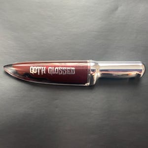 May include: A silver and burgundy lip gloss tube shaped like a knife with the words "GOTH GLOSSED" printed on the side.