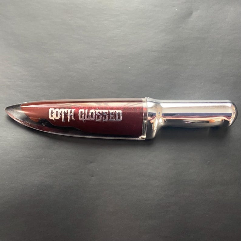 May include: A silver and burgundy lip gloss tube shaped like a knife with the words "GOTH GLOSSED" printed on the side.