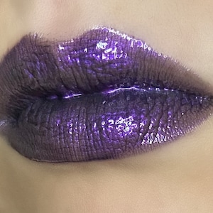 May include: Close-up of a person's lips wearing a dark purple lip gloss with a shimmery finish.