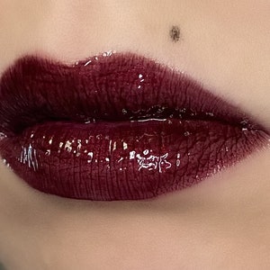 May include: Close-up of a person's lips wearing a dark burgundy lip gloss with a glossy finish.