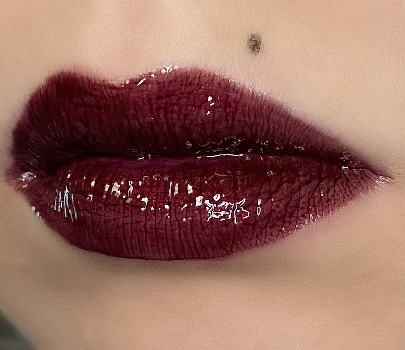 May include: Close-up of a person's lips wearing a dark burgundy lip gloss with a glossy finish.