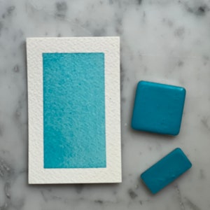 Puede incluir: Una muestra de acuarela sobre papel texturizado con un bloque de pintura rectangular turquesa. También hay dos objetos rectangulares turquesa. Los objetos se muestran sobre una superficie de mármol. La imagen muestra materiales de arte y muestras de color.