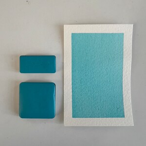 Peut inclure: Deux petits pots de peinture aquarelle bleu sarcelle à côté d'un échantillon plus grand de la même couleur peint sur du papier aquarelle.