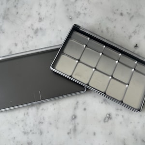 Puede incluir: Una paleta de pintura de metal plateado con 12 huecos vacíos. La paleta está abierta y la tapa está puesta al lado.