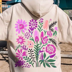 May include: A beige hoodie with a floral design on the back. The flowers are pink, purple, and green. The hoodie has a hood and a kangaroo pocket.