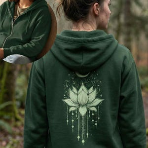 May include: Dark green zip-up hoodie with a hood. The back of the hoodie features a white lotus flower design with a crescent moon and star accents. The hoodie is made of a soft material.
