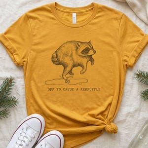 May include: A mustard yellow t-shirt featuring a black line art illustration of a raccoon standing on its hind legs. Below the raccoon is a small puddle and the text "OFF TO CAUSE A KERFUFFLE" printed in a simple, dark, sans-serif font.