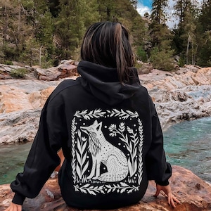May include: Black hooded sweatshirt with a white graphic print of a fox in a floral frame. The fox is sitting and looking to the right. The sweatshirt is worn by a person standing in front of a rocky riverbed.