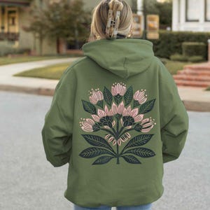 May include: An olive green hoodie featuring a floral design on the back. The design includes pink flowers, dark green leaves, and stems. The hoodie has a hood and a relaxed fit, suitable for casual wear.