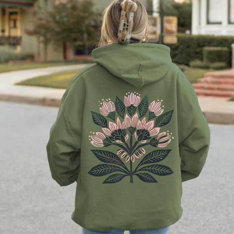 May include: An olive green hoodie featuring a floral design on the back. The design includes pink flowers, dark green leaves, and stems. The hoodie has a hood and a relaxed fit, suitable for casual wear.
