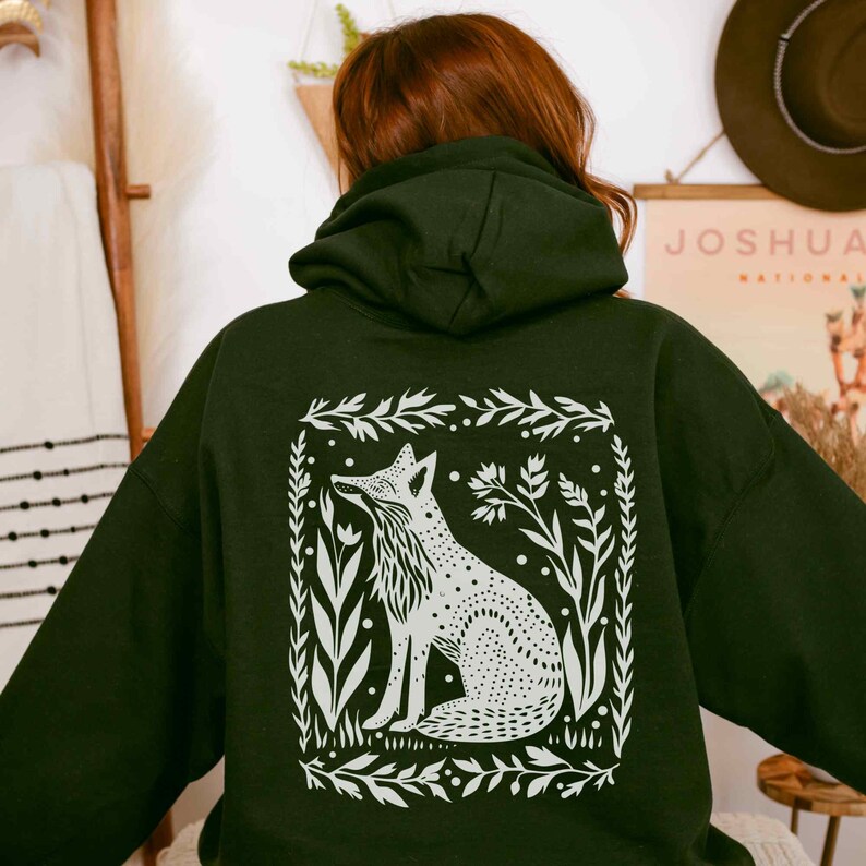 May include: A black hooded sweatshirt with a white fox illustration on the back. The fox is surrounded by a leafy border and is sitting in a field of flowers.
