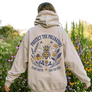 Peut inclure: Un sweat-shirt à capuche beige avec un motif au dos représentant une abeille centrale entourée de fleurs jaunes et bleues. Le texte indique "PROTECT THE POLLINATORS" en arc au-dessus et "PLANT NATIVE SAVE THE BEES" en bas.