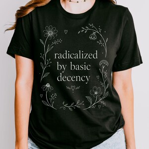May include: A black short-sleeved t-shirt featuring white line art of a floral wreath surrounding the text "radicalized by basic decency" in a serif font. The design includes small heart icons interspersed among the flowers and stems.
