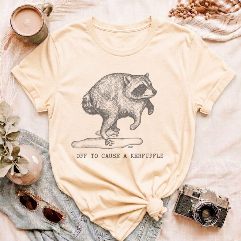 May include: A light yellow short-sleeve t-shirt featuring a dark gray line art illustration of a raccoon standing on a puddle with a paw print. Below the illustration, the text reads OFF TO CAUSE A KERFUFFLE in a simple, dark gray sans-serif font.