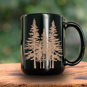 May include: Black ceramic coffee mug featuring a tan illustration of several pine trees. The mug has a large handle and is sitting on a wooden surface. The background is blurred, suggesting an outdoor setting.