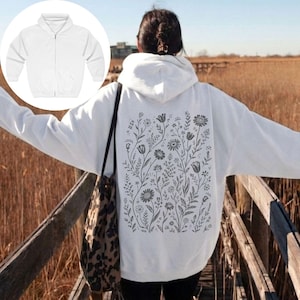 May include: A white zip-up hoodie featuring a large, dark grey botanical line art illustration on the back, depicting a variety of wildflowers, stems, and leaves. The hoodie includes a hood, long sleeves, and a front zip closure design.