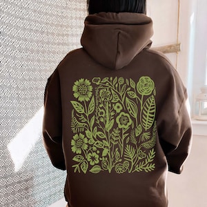 May include: A brown hoodie with a green floral design on the back. The design features a variety of flowers, leaves, and stems.