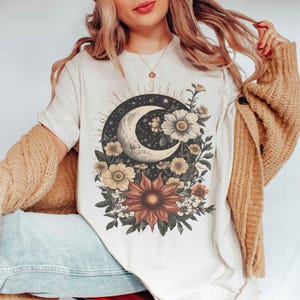 Może przedstawiać: Kremowa koszulka z niebiańskim wzorem. Półksiężyc otoczony jest ciemnym okręgiem z gwiazdami i elementami kwiatowymi, w tym słonecznikami i stokrotkami w odcieniach brązu, żółci i bieli. Koszulka ma krótkie rękawy.