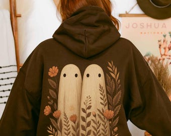 May include: A dark brown hooded sweatshirt featuring a back graphic of two beige ghost figures standing side by side, surrounded by various illustrated botanical elements including flowers and leaves in shades of orange and brown.