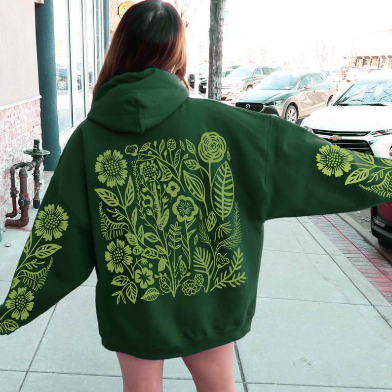 Puede incluir: Sudadera con capucha verde oscuro con un dise&ntilde;o floral en verde claro. El dise&ntilde;o presenta varias flores, hojas y tallos, cubriendo la espalda y las mangas. La sudadera tiene capucha y un corte holgado.