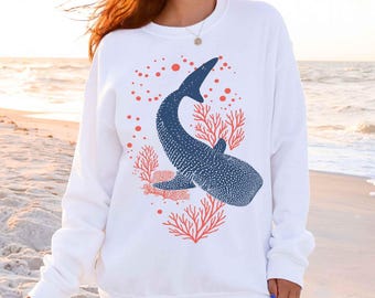 May include: A white long-sleeve crewneck sweatshirt featuring a dark blue whale shark graphic surrounded by orange coral and bubble illustrations. The design is centered on the chest, showcasing the spotted pattern of the whale shark.