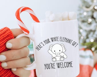May include: A white ceramic mug filled with white marshmallows and a red and white striped candy cane. The mug features a black line drawing of a baby elephant and the text: HERE'S YOUR WHITE ELEPHANT GIFT YOU'RE WELCOME printed in black.