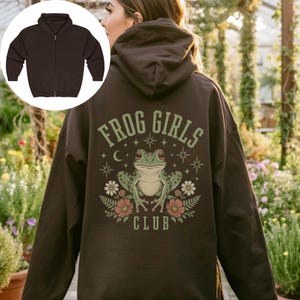 May include: A dark brown zip-up hoodie featuring a back graphic of a green frog surrounded by flowers, stars, and a crescent moon. The text "FROG GIRLS CLUB" is printed in a light green arched font above and below the central frog illustration.