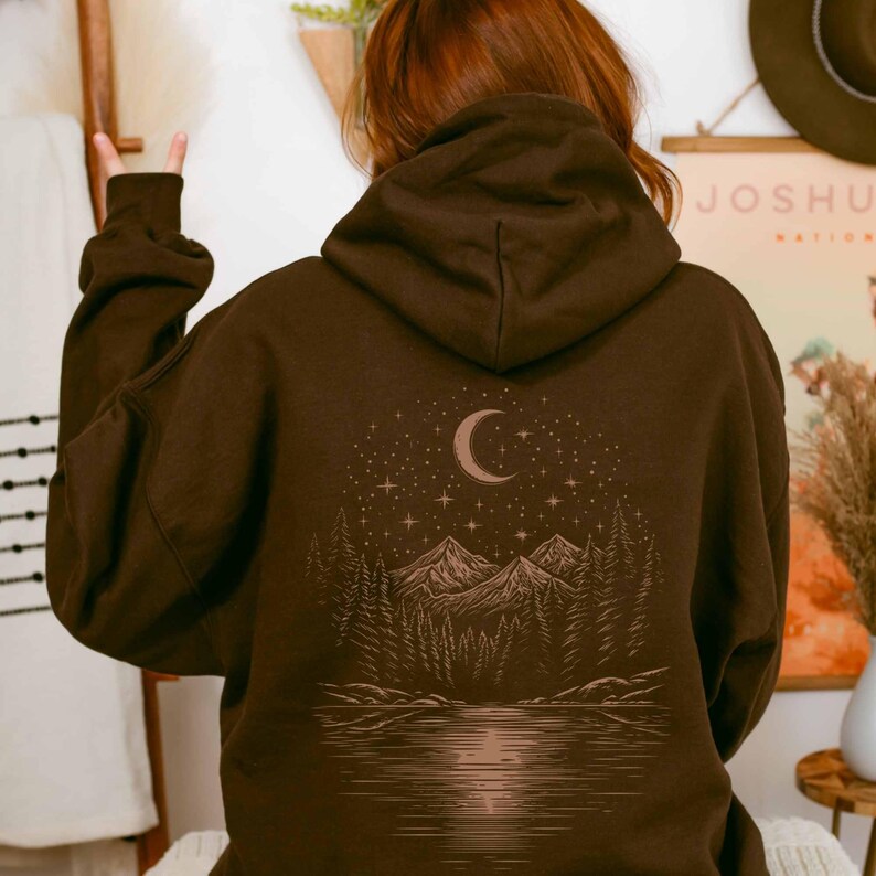 May include: A brown hoodie with a nature-themed graphic on the back. The design features a crescent moon, stars, mountains, trees, and a lake. The hoodie has a hood and long sleeves.