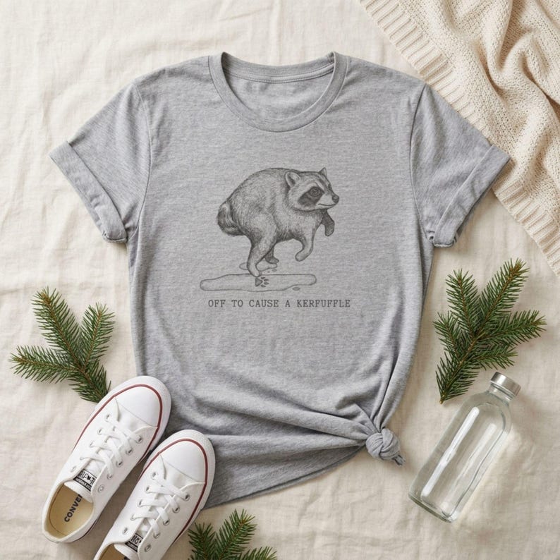 May include: A heather gray short-sleeve t-shirt featuring a black line-art illustration of a raccoon stepping into a puddle. Below the raccoon, the text reads: OFF TO CAUSE A KERFUFFLE. The shirt has a crew neckline and rolled sleeves.