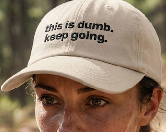 May include: A beige baseball cap featuring the embroidered black text "this is dumb. keep going." on the front panel. The cap has a structured crown, a curved brim, and stitched eyelets on the top panels for ventilation and a classic fit.