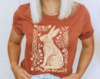 May include: A rust-colored short-sleeve t-shirt featuring a cream-colored graphic print of a rabbit sitting in profile, surrounded by a rectangular frame of stylized leaves and small flowers. The shirt has a crew neckline and a heathered texture.