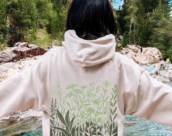 May include: A beige hooded sweatshirt featuring a large rectangular graphic on the back. The print displays a detailed botanical illustration of various wildflowers and grasses in shades of light and dark sage green against the fabric.